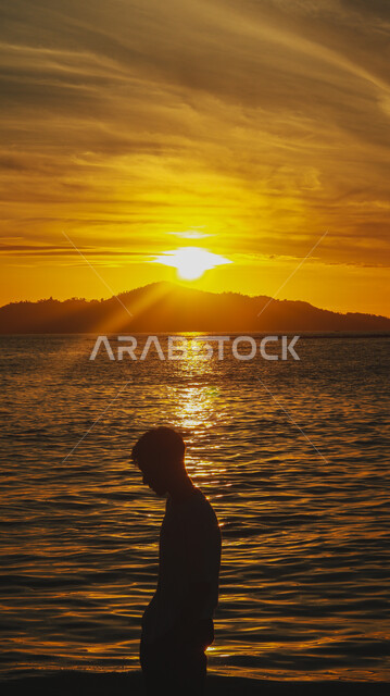 A view of the sunset over the sea coast, areas for trips and picnics in the summer, spending a pleasant time contemplating the tranquil landscape, a silhouette from behind of a young Saudi Arabian Gulf man standing on the sandy beach, natural places and the waterfront in the Kingdom of Saudi Arabia, Silhouette
