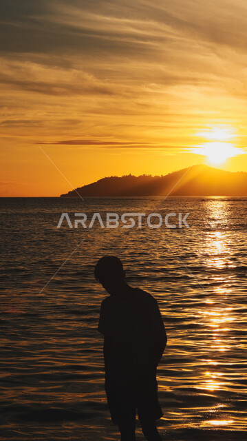 مشهد لغروب الشمس فوق ساحل البحر، مناطق الرحلات والنزهات في الصيف، قضاء وقت ممتع بتأمل مناظر الطبيعة الهادئة، صورة ظلية من الخلف لشاب عربي خليجي سعودي يقف على الشاطئ الرملي، الأماكن الطبيعية والواجهة البحرية في المملكة العربية السعودية، سالويت  