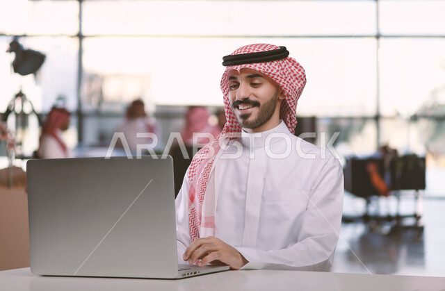 مهن ووظائف ادارية مكتبية، دمج العمل مع التقنية والتكنولوجيا، إدارة شؤون الأعمال وتنظيمها عبر الاجهزة التقنية، رجل اعمال عربي خليجي سعودي يرتدي الشماغ والثوب التقليدي عمل على الحاسوب المحمول داخل المكتب