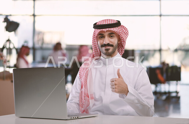 إدارة شؤون الأعمال وتنظيمها عبر الاجهزة التقنية، مهن ووظائف ادارية مكتبية، دمج العمل مع التقنية والتكنولوجيا، رجل اعمال عربي خليجي سعودي يرتدي الشماغ والثوب التقليدي عمل على الحاسوب المحمول داخل المكتب يرفع يده بايماءات الجودة