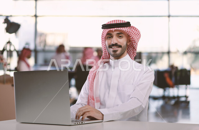 Integrating work with technology and technology, office administrative professions and jobs, managing and organizing business affairs through technical devices, a Saudi Arabian Gulf businessman wearing a shemagh and traditional thobe working on a laptop inside the office.