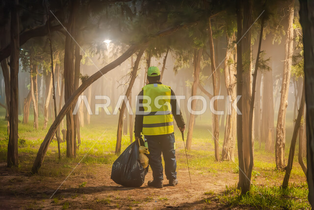 الاهتمام بنظافة البيئة، صورة من الخلف لعامل نظافة عربي خليجي سعودي يرتدي الزي الخاص بالعمل يحمل بيده كيس اسود، إزالة النفايات والقمامة من الحدائق المنتزهات العامة، حديقة ابها المليئة بالاشجار والنباتات الخضراء