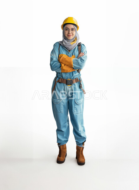 العمل في القطاع الهندسي السعودي، مهن ووظائف هندسية نسائية، بورتريه لفتاة عربية خليجية سعودية ترتدي الخوذة وزي العمل تقف بأيدي متقاطعة، تعابير وايماءات الثقة بالنفس والسرور، صورة الجسم بالطول الكامل، خلفية بيضاء