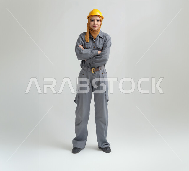 مهن ووظائف هندسية نسائية، بورتريه لفتاة عربية خليجية سعودية ترتدي الخوذة وزي العمل تقف بأيدي متقاطعة، تعابير وايماءات الثقة بالنفس والسرور، العمل في القطاع الهندسي السعودي، صورة الجسم بالطول الكامل، خلفية بيضاء