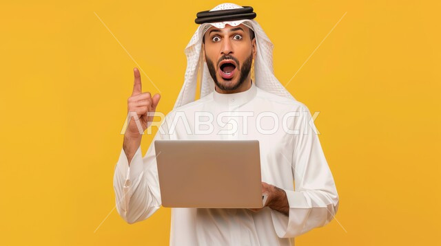 التفاجؤ من شيء ما، استخدام الأجهزة والتقنيات الحديثة، الشعور بالدهشة والتعجب والصدمة والذهول , بورتريه مقرب لشاب عربي خليجي سعودي يرتدي الشماغ والزي التقليدي يمسك بيده الحاسوب المحمول يشير بإصبع السبابة إلى شيء ما، الإشارة إلى مكان ما، خلفية صفراء