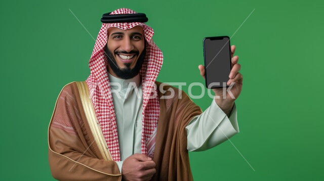 عرض شاشة الهاتف المحمول، بورتريه لرجل عربي خليجي سعودي مبتسم يرتدي الشماغ والثوب التقليدي يمسك بيده الجوال، النظر إلى الكاميرا بإيماءات السرور والسعادة، الاهتمام بالمظهر الخارجي, خلفية خضراء