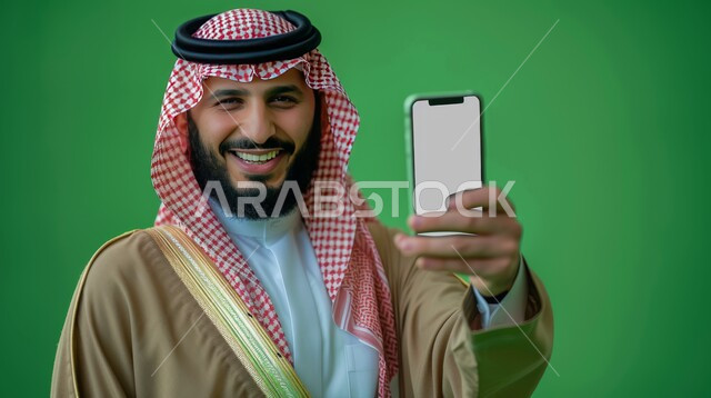 Paying attention to external appearance, looking at the camera with gestures of pleasure and happiness, displaying a mobile phone screen, a portrait of a smiling Saudi Arabian Gulf man wearing a shemagh and traditional thobe, holding a mobile phone in his hand, green background
