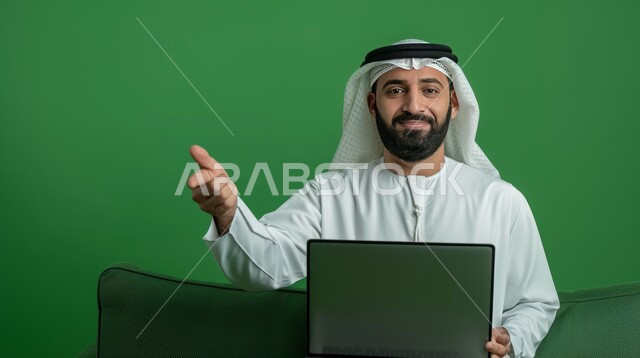 استخدام اللابتوب والعمل عن بعد، بورتريه مقرب لرجل عربي خليجي سعودي مبتسم يرتدي الشماغ والثوب التقليدي يحمل الحاسوب المحمول يشير بإصبع السبابة إلى شيء ما، خلفية خضراء