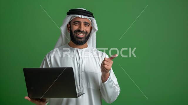 الإشارة بإصبع السبابة إلى شيء ما، استخدام اللابتوب والعمل عن بعد، بورتريه مقرب لرجل عربي خليجي سعودي مبتسم يرتدي الشماغ والثوب التقليدي يحمل الحاسوب المحمول، خلفية خضراء