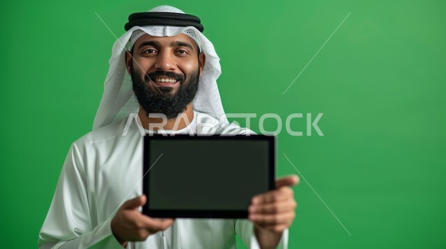 Displaying the personal computer screen, looking at the camera with happy expressions, using the laptop and working remotely, a close-up portrait of a smiling Saudi Gulf Arab man wearing a shemagh and traditional thobe, holding the laptop, green background