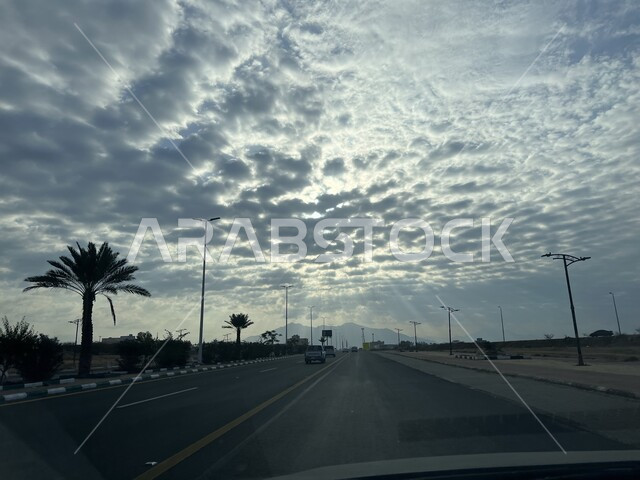 حركة سير السيارات في الطرقات والشوارع الاسفلتية المعبدة، أشجار النخيل الخضراء على الطريق السريع في مدينة العيدابي بالمملكة العربية السعودية، السماء المليئة بالغيوم فوق شارع مجهز بأعمدة الإنارة الطرقية
