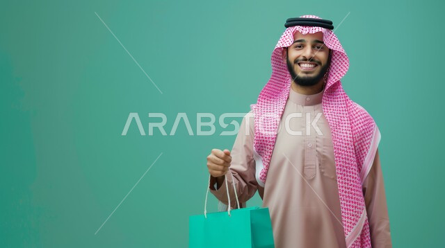 Expressions and movements of enthusiasm for shopping and purchasing supplies and gifts, taking advantage of Black Friday offers and sales, buying items and the joy of shopping in malls and shops, a close-up portrait of a smiling Saudi Arabian Gulf man wearing a shemagh and traditional thobe, holding colored paper bags in his hand, blue background.