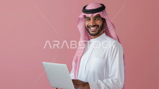 استخدام اللابتوب والعمل عن بعد، النظر إلى الكاميرا بتعابير السرور، بورتريه مقرب لرجل عربي خليجي سعودي مبتسم يرتدي الشماغ والثوب التقليدي يحمل الحاسوب المحمول، خلفية وردية
