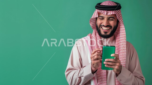 عرض شاشة الهاتف المحمول، إيماءات السرور والسعادة، بورتريه مقرب لرجل عربي خليجي سعودي مبتسم يرتدي الشماغ والثوب التقليدي يمسك بيده الجوال، الاهتمام بالمظهر الخارجي, خلفية خضراء