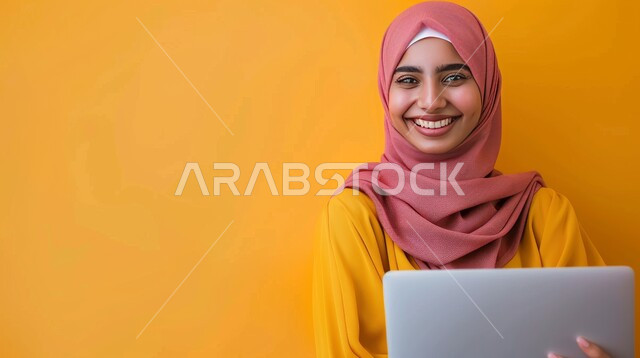 استخدام الوسائل التقنية الحديثة والمتطورة لإنجاز المهام، النظر إلى الكاميرا بتعابير السرور، بورتريه مقرب لإمرأة عربية خليجية سعودية مبتسمة محجبة تحمل بيدها الحاسوب المحمول تعمل على الحاسوب المحمول، خلفية صفراء