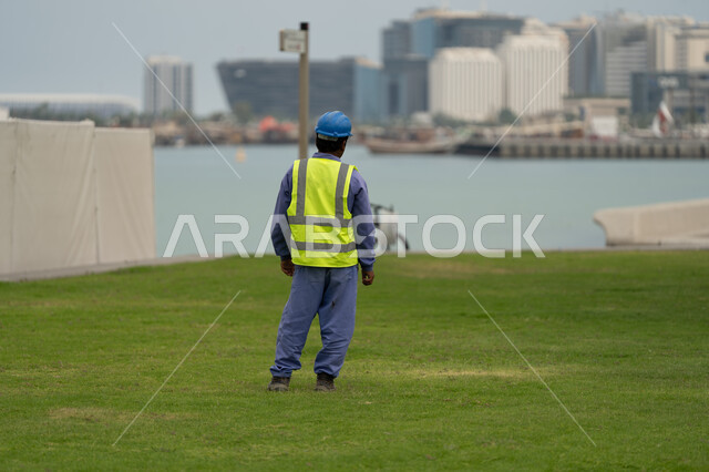 النمو والتقدم العمراني للأبراج والمباني المطلة على البحر الأحمر، صورة من الخلف لعامل يرتدي زي العمل وخوذة الحماية يقوم بقضاء وقت الإستراحة على كورنيش مدينة الدوحة، الأماكن والمعالم الساحلية السياحية في دولة قطر