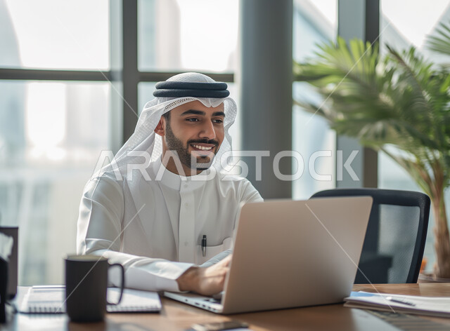 دمج العمل مع التقنية والتكنولوجيا، مهن ووظائف ادارية مكتبية، إدارة شؤون الأعمال وتنظيمها عبر الاجهزة التقنية، رجل اعمال عربي خليجي سعودي يرتدي الزي التقليدي والغترة يعمل على الحاسوب المحمول داخل المكتب