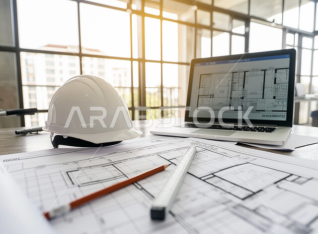 Saudi engineering professions and jobs, white safety helmet on the desk, equipment, tools and building plans on an office table, following up on work and designs via the laptop, completing work tasks and drawing projects using modern technologies.