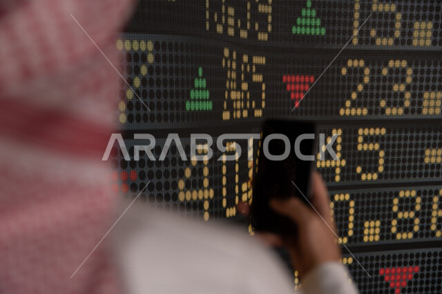 رجل اعمال سعودي خليجي يستخدم البورصة و الاسهم لتطوير اعماله ، يحمل بيده هاتفه المحمول  ، استخدام تقنية الجرافيكس في عرض نزول و صعود الاسهم  ، أخبار عالمية اقتصادية ،نشرة الإحصاءات الاقتصادية للدول العربية