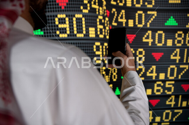 رجل اعمال سعودي خليجي يستخدم البورصة و الاسهم لتطوير اعماله ، يحمل بيده هاتفه المحمول  ، استخدام تقنية الجرافيكس في عرض نزول و صعود الاسهم  ، أخبار عالمية اقتصادية ،نشرة الإحصاءات الاقتصادية للدول العربية