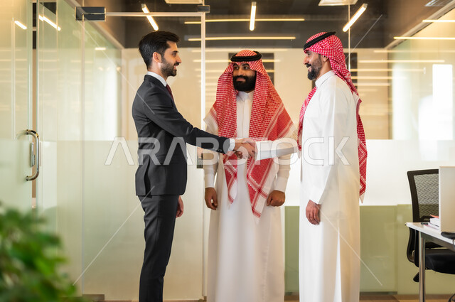 الاتفاق على المشاريع التجارية، رجل عربي خليجي سعودي يرتدي الثوب والشماغ التقليدي يصافح رجل اعمال يرتدي البدلة الرسمية، فريق عمل داخل غرفة الاجتماعات في مقر الشركة، الترحيب والسلام وإلقاء التحية