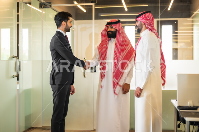 الاتفاق على المشاريع التجارية، رجل عربي خليجي سعودي يرتدي الثوب والشماغ التقليدي يصافح رجل اعمال يرتدي البدلة الرسمية، فريق عمل داخل غرفة الاجتماعات في مقر الشركة، الترحيب والسلام وإلقاء التحية