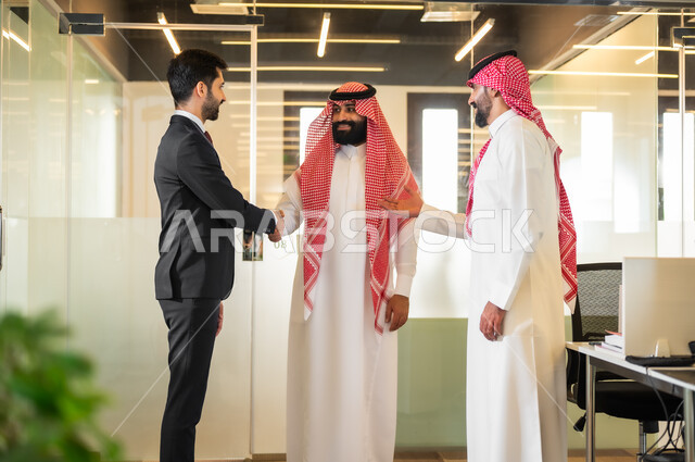 الاتفاق على المشاريع التجارية، رجل عربي خليجي سعودي يرتدي الثوب والشماغ التقليدي يصافح رجل اعمال يرتدي البدلة الرسمية، فريق عمل داخل غرفة الاجتماعات في مقر الشركة، الترحيب والسلام وإلقاء التحية