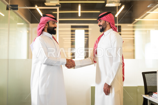 Welcoming, greeting, greetings, two smiling Saudi Gulf Arab men wearing the traditional shemagh and thobe, shaking hands with gestures of pleasure at the success of the deal, unity and group interaction to achieve the company’s goals and solve problems, professions and office jobs.