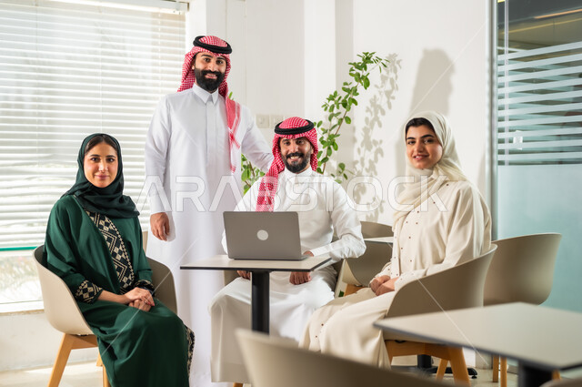 النظر الى الكاميرا بايماءات السعادة والسرور لبدء الشراكة، اجتماع عمل بين موظفين عرب خليجيين سعوديين في غرفة الاجتماعات، التفاهم والتعاون بين الزملاء لتحقيق أهداف الشركة, بيئة العمل المريحة في الشركات السعودية, استخدام الأجهزة التقنية الحديثة في المكاتب