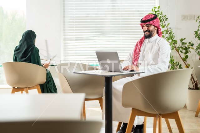 وظائف ومهن إدارية سعودية، إدارة شؤون العمل وتنظيمها، موظف عربي خليجي سعودي يرتدي الزي التقليدي يجلس أمام الحاسوب المحمول، موظفة تقوم بتدقيق أوراق العمل، دمج التقنيات الحديثة والمتطورة بالأعمال المكتبية، بيئة العمل المريحة في الشركات السعودية