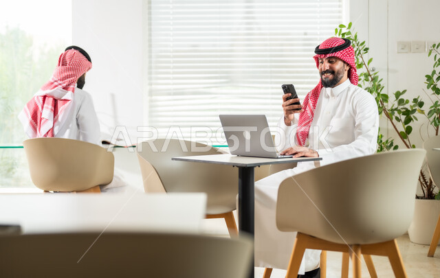 إدارة شؤون العمل وتنظيمها، وظائف ومهن إدارية سعودية، موظف عربي خليجي سعودي يرتدي الزي التقليدي يجلس أمام الحاسوب المحمول وينظر الى الهاتف، شاب يقوم بتدقيق أوراق العمل، دمج التقنيات الحديثة والمتطورة بالأعمال المكتبية، بيئة العمل المريحة في الشركات السعودية