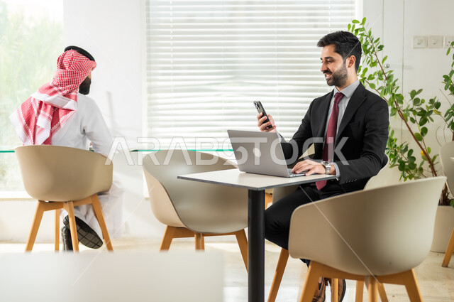 بيئة العمل المريحة في الشركات السعودية، دمج التقنيات الحديثة بالأعمال المكتبية، إدارة شؤون العمل وتنظيمها، وظائف ومهن إدارية سعودية، موظف يجلس أمام الحاسوب ويقوم بالنظر الى الهاتف، شاب عربي خليجي سعودي يرتدي الزي التقليدي يقوم بتدقيق أوراق العمل