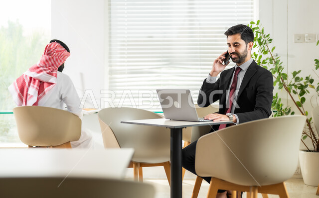 Using modern technologies in office work, managing and organizing work affairs, Saudi administrative jobs and professions, an employee sitting in front of the computer and making a phone call, a Saudi Gulf Arab young man wearing traditional clothing checking work papers, a comfortable work environment in Saudi companies