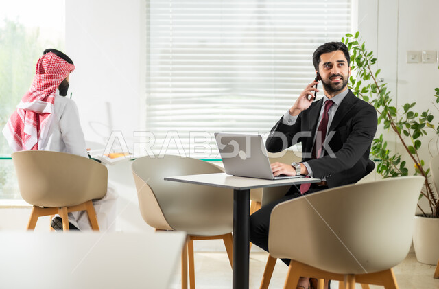 دمج التقنيات الحديثة بالأعمال المكتبية، إدارة شؤون العمل وتنظيمها، وظائف ومهن إدارية سعودية، موظف يجلس أمام الحاسوب ويقوم باجراء مكالمة هاتفية، شاب عربي خليجي سعودي يرتدي الزي التقليدي يقوم بتدقيق أوراق العمل، بيئة العمل المريحة في الشركات السعودية