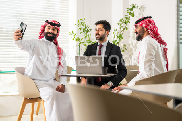 انجاز مهام العمل باستخدام جهاز اللابتوب، التقاط صور تذكارية عبر الموبايل، مهن ووظائف مكتبية سعودية، إجتماع عمل لمناقشة أهداف الشركة, صفقات وإتفاقيات عمل جديده، رجلا أعمال عربيان خليجيان سعوديان يرتديان الزي التقليدي مع موظف باللباس الرسمي في غرفة الإجتماعات