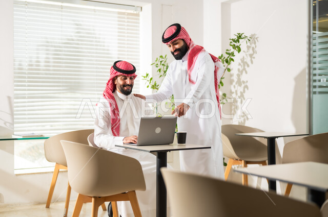 مناقشة المشاريع ومتابعة إستراتيجية العمل، موظفان عربيان خليجيان سعوديان يرتديان الزي التقليدي يقومان بمراجعة البحوث العلمية، إستخدام الحاسوب المحمول لإنجاز المهام، مهن ووظائف مكتبية سعودية، مفهوم إدارة الأعمال، تحقيق أهداف الشركة