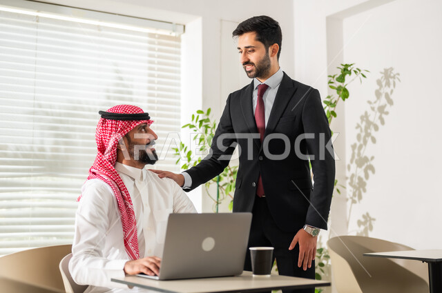 مفهوم إدارة الأعمال، إنجاز المهام عبر الحاسوب المحمول، مناقشة المشاريع ومتابعة إستراتيجية العمل، موظفان عربيان خليجيان سعوديان يرتديان الزي التقليدي يقومان بمراجعة البحوث العلمية، مهن ووظائف مكتبية سعودية، تحقيق أهداف الشركة