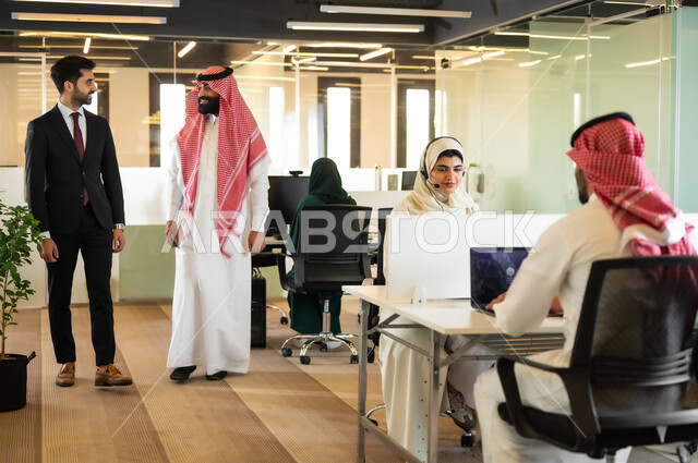 مناقشة الخطط وشرح مهام الاعمال، رجل عربي سعودي خليجي يرتدي الثوب التقليدي والشماغ مع  رجل بالبدلة الرسمية يقفون داخل مقر الشركة، الاتفاق على المشاريع التجارية، بيئة العمل المريحة في الشركات السعودية, حوارات و مناقشات لتحقيق الاستراتيجيات والغايات