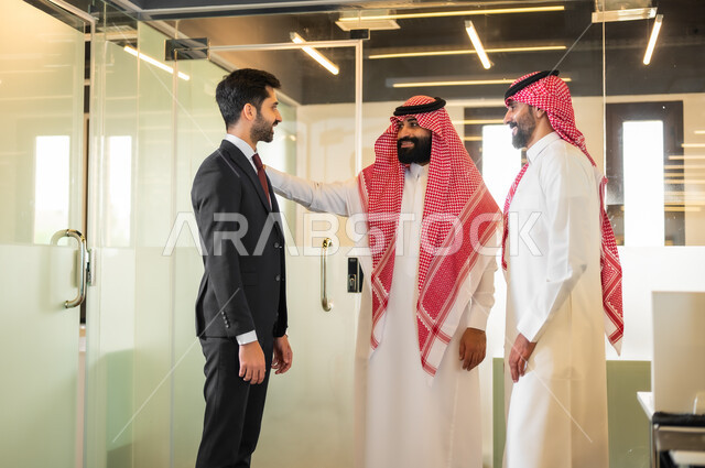 Discussing plans and explaining business tasks, the comfortable work environment in Saudi companies, two Saudi Gulf Arab men wearing the traditional thobe and shemagh with a man in a formal suit standing inside the company headquarters, agreeing on business projects, dialogues and discussions to achieve strategies and objectives.