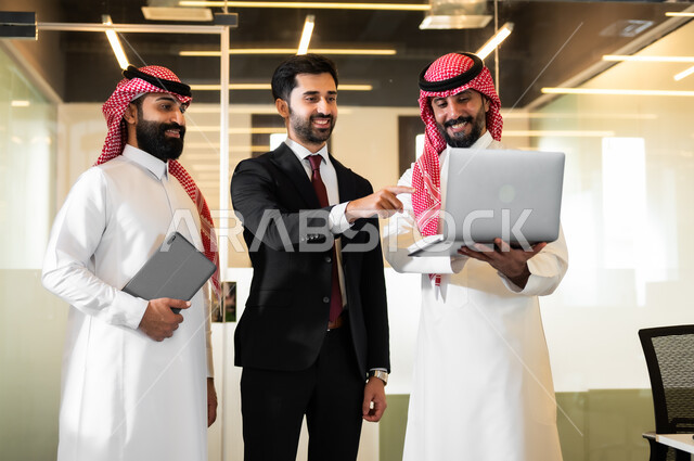 متابعة ومناقشة إستراتيجيات وأهداف الشركة، استخدام الاجهزة والتقنيات الحديثة في تطوير الاعمال، رجلان عربيان سعوديان خليجيان يرتديان الثوب التقليدي والشماغ مع رجل بالبدلة الرسمية يقفون داخل مقر الشركة، الاشارة الى الحاسوب بهدف توضيح المعلومات