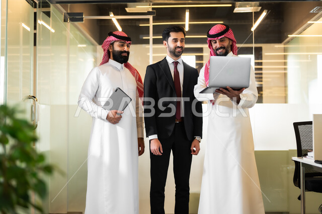 مناقشة إستراتيجيات وأهداف الشركة، استخدام الاجهزة والتقنيات الحديثة في تطوير الاعمال، رجلان عربيان سعوديان خليجيان يرتديان الثوب التقليدي والشماغ مع رجل بالبدلة الرسمية يقفون داخل مقر الشركة، الاشارة الى الحاسوب بهدف توضيح المعلومات