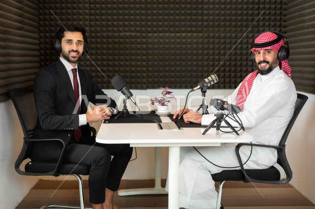 النظر إلى الكاميرا بايماءات السرور، رجل عربي خليجي سعودي يرتدي الثوب والشماغ التقليدي مع رجل بالبدلة الرسمية في مقابلة عمل داخل مقر الشركة، معدات وأجهزة تسجيلات الصوت،  اجراء بث صوتي مباشر ( بودكاست ) عبر برامج التواصل الاجتماعي