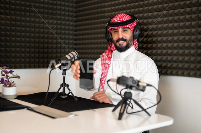 Showing a blank black screen, a smiling Saudi Gulf Arab man wearing a traditional thobe and shemagh in a job interview inside the company headquarters looking at the camera, gestures of happiness and pleasure, audio recording equipment and devices, conducting a live audio broadcast (podcast) via social media programs