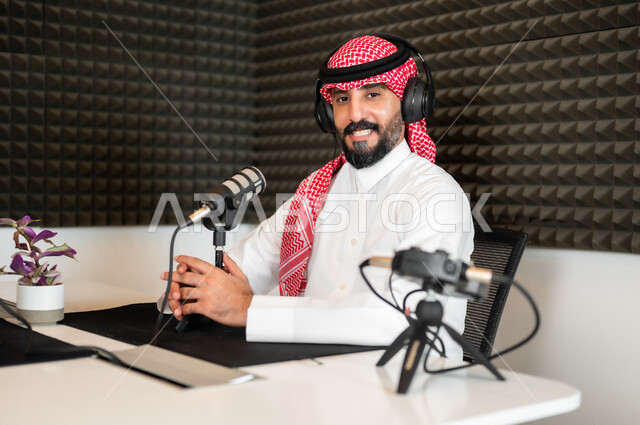 النظر إلى الكاميرا بإيماءات السرور، رجل عربي خليجي سعودي مبتسم يرتدي الثوب والشماغ التقليدي في مقابلة عمل داخل مقر الشركة، معدات وأجهزة تسجيلات الصوت،  اجراء بث صوتي مباشر ( بودكاست ) عبر برامج التواصل الاجتماعي
