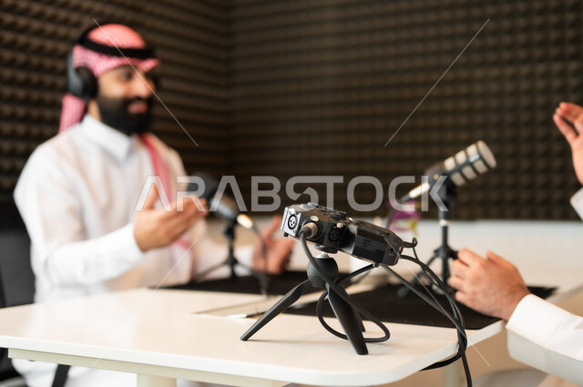 Audio recording studio, conducting a live broadcast (podcast) via social media programs, a smiling Saudi Gulf Arab man wearing the traditional thobe and shemagh, raising his hands up, audio recording equipment and devices, gestures of enthusiasm and pleasure.