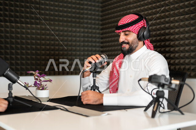 ايماءات السعادة والسرور، رجل عربي خليجي سعودي مبتسم يرتدي الثوب والشماغ التقليدي في مقابلة عمل داخل مقر الشركة، معدات وأجهزة تسجيلات الصوت،  اجراء بث صوتي مباشر ( بودكاست ) عبر برامج التواصل الاجتماعي