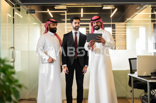 مناقشة مهام العمل،  بيئة العمل المريحة في الشركات السعودية، رجلان عربيان سعوديان خليجيان يرتديان الثوب التقليدي والشماغ مع رجل بالبدلة الرسمية يقفون داخل مقر الشركة، الاتفاق على المشاريع التجارية، إنجاز المهام العملية عبر جهاز التابليت