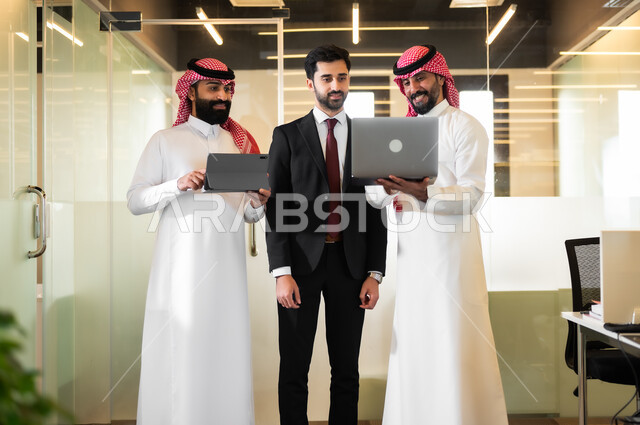 متابعة ومناقشة إستراتيجيات وأهداف الشركة، استخدام الاجهزة والتقنيات الحديثة في تطوير الاعمال، رجلان عربيان سعوديان خليجيان يرتديان الثوب التقليدي والشماغ مع رجل بالبدلة الرسمية يقفون داخل مقر الشركة، الاشارة الى الحاسوب بهدف توضيح المعلومات