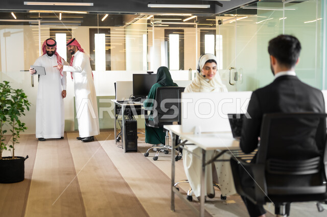 استخدام الأجهزة والتقنيات الحديثة لتسهيل الأعمال، رجلان عربيان سعوديان خليجيان يرتديان الثوب التقليدي والشماغ يقفون داخل مقر الشركة، الوحدة والتفاعل الجماعي لتحقيق أهداف الشركة وحل المشكلات، مهن و وظائف مكتبية