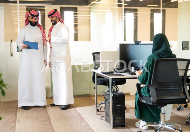 Work talks and discussions, unity and group interaction to achieve the company’s goals, using modern devices and technologies to facilitate work, two Saudi and Gulf Arab men wearing the traditional thobe and shemagh, completing practical tasks via a tablet.
